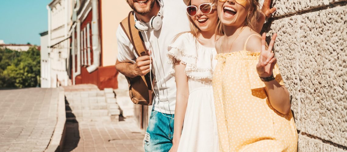 Group of young three stylish friends posing in the street. Fashion man and two cute girls dressed in casual summer clothes. Smiling models having fun in sunglasses.Cheerful women and guy outdoors